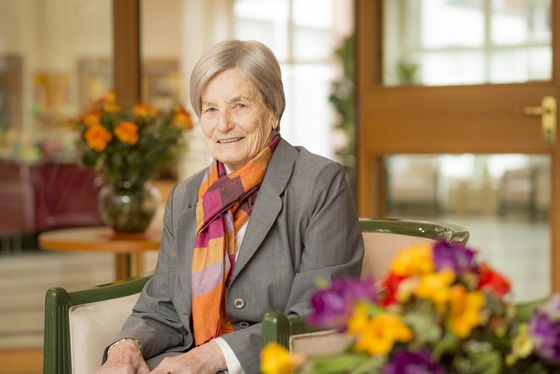 Eine Seniorin sitzt lächelnd auf einem Stuhl vor einem mit Blumen dekorierten Tisch, pflegerische Unterstützung im Ausnahmefall, Kurzzeithilfe nach einem Krankenhausaufenthalt, temporäre Pflege, Residenz am Wiesenkamp, Hamburg-Volksdorf Eine Seniorin sitzt lächelnd auf einem Stuhl vor einem mit Blumen dekorierten Tisch, pflegerische Unterstützung im Ausnahmefall, Kurzzeithilfe nach einem Krankenhausaufenthalt, temporäre Pflege, Residenz am Wiesenkamp, Hamburg-Volksdorf