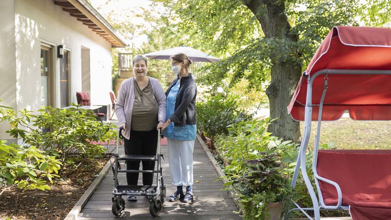 Diakonie Hospiz Wannsee - Eine Pflegerin begleitet eine Hospizgästin auf die von einem Park umgebene Terrasse des Diakonie Hospiz Wannsee. 