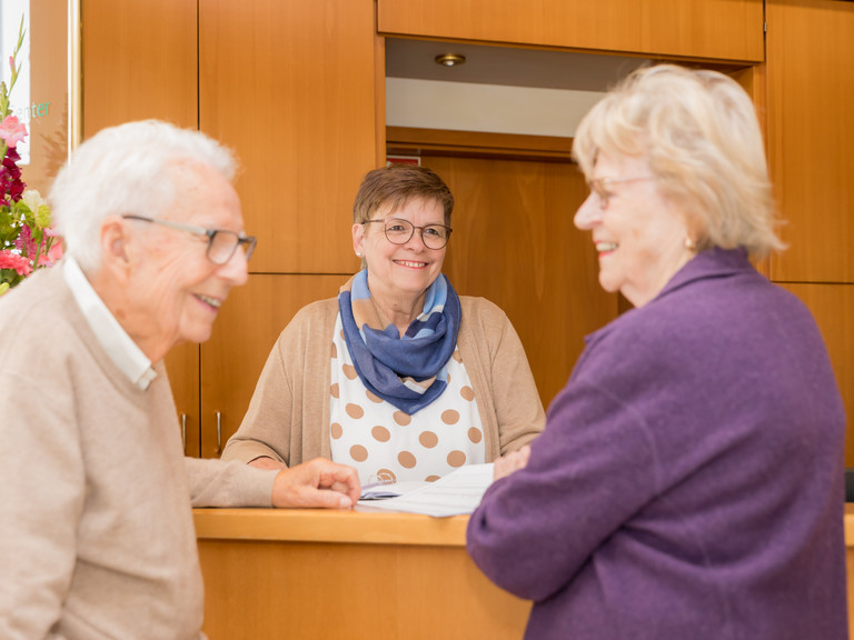 Eine Seniorin und ein Senior werden an der Rezeption von einer Mitarbeiterin beraten, Leistungen der Residenz am Wiesenkamp, Hamburg-Volksdorf