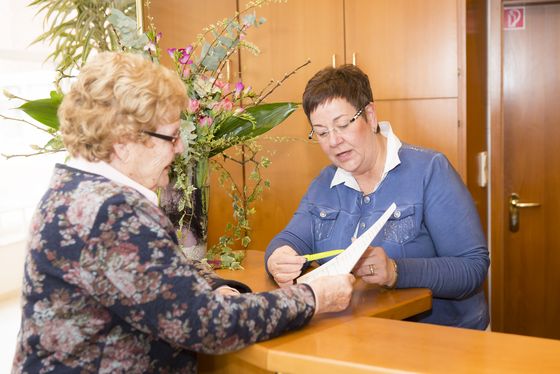 Eine Seniorin wird an der Rezeption von einer Mitarbeiterin bedient, enthaltene Leistungen im Wohngeld, Unterstützung im Alltag im Seniorenheim, Angebot in der Residenz am Wiesenkamp, Hamburg-Volksdorf Eine Seniorin wird an der Rezeption von einer Mitarbeiterin bedient, enthaltene Leistungen im Wohngeld, Unterstützung im Alltag im Seniorenheim, Angebot in der Residenz am Wiesenkamp, Hamburg-Volksdorf